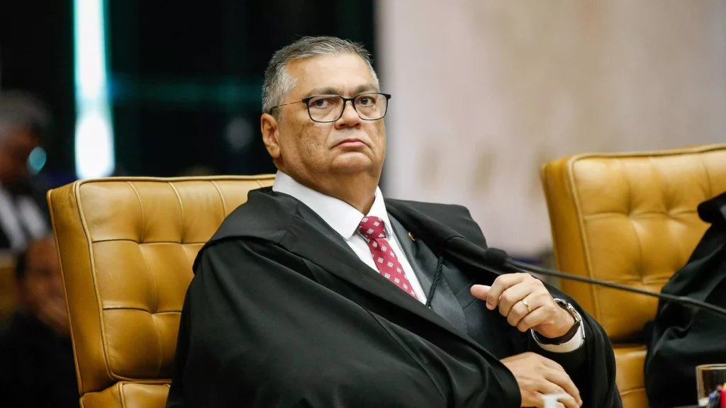 Ministro do STF Flávio Dino sentado em uma cadeira de couro caramelo durante sessão, usando toga preta, terno, gravata vermelha estampada e óculos, com expressão séria.