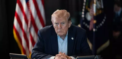 Donald Trump, presidente dos Estados Unidos, sentado à mesa diante de bandeiras americanas.