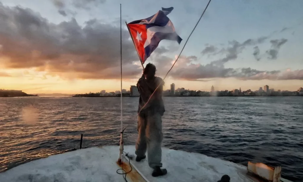 Um homem hasteia a bandeira de Cuba em um barco, com o pôr do sol e a skyline de Havana ao fundo. (Foto: nodal.am)
