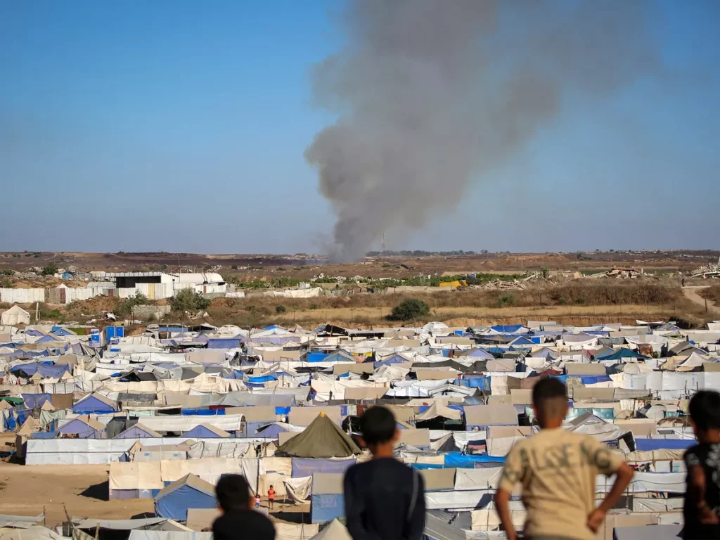 Crianças observam colunas de fumaça em campo de refugiados em Gaza. (Foto: aljazeera.com)