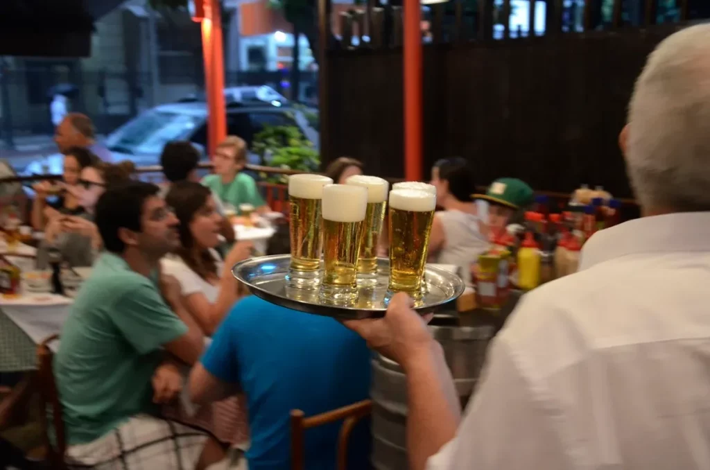 Garçom serve cervejas em um bar movimentado à noite. (Foto: https://www.ocafezinho.com/2026/04/17/vida-noturna-no-brasil-um-g
