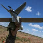 Soldado carrega drone em campo com flores vermelhas sob céu azul. (Foto: rt.com)