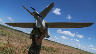 Soldado carrega drone em campo com flores vermelhas sob céu azul. (Foto: rt.com)