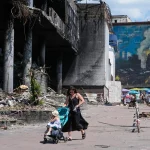 Mulher empurra carrinho com criança em rua destruída por bombardeios em cidade ucraniana. (Foto: aljazeera.com)