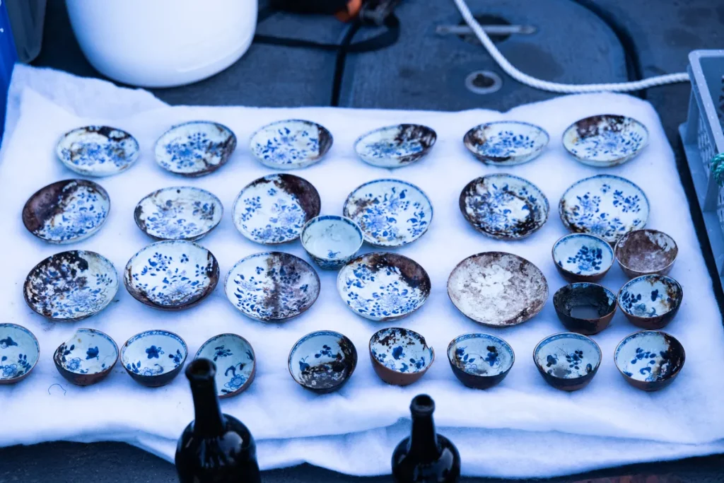 Várias peças de porcelana chinesa com desenhos azuis e brancos são exibidas sobre tecido branco, possivelmente após recuperaç