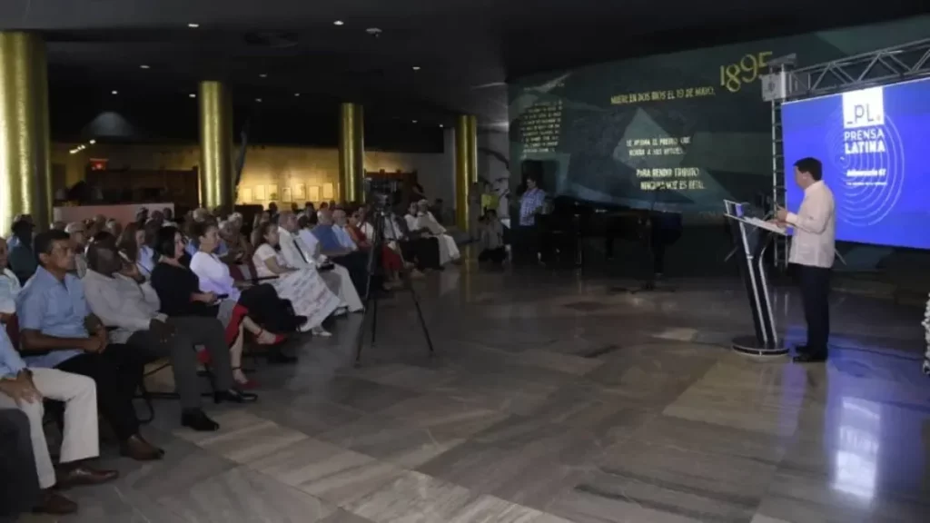 Um homem discursa em evento comemorativo da Agencia Prensa Latina, com público atento e telão ao fundo. (Foto: telesurtv.net)
