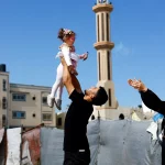 Um homem levanta uma criança em Gaza, com uma mesquita ao fundo. (Foto: aljazeera.com)