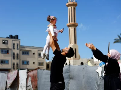 Um homem levanta uma criança em Gaza, com uma mesquita ao fundo. (Foto: aljazeera.com)